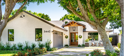 Modern white brick house with grey double garage and wood doors. Chandelier hangs over the open porch under a blue sky, trees cover the drive