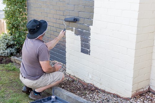 Man crouched using a roller to paint a white exterior brick wall grey