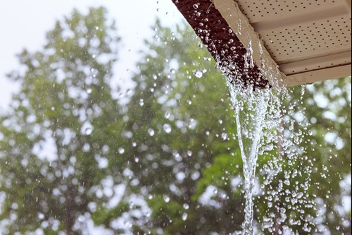 View from under a roof corner with rainwater spilling over the edge of a gutter, trees visible in the background