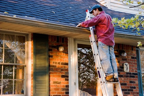 Man in a red shirt and ball cap standing on a ladder, removing debris from the gutter of a red brick house