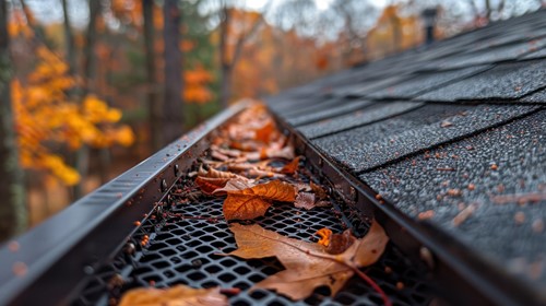 Close-up of a mesh gutter guard blocking fall leaves from entering the gutter, with orange autumn trees in the background