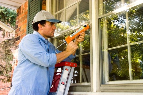 Man on a ladder using a caulking gun to seal the exterior windows of a brick house