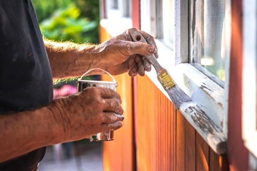 Man painting white window trim on a red house using a small brush, holding a can of white paint