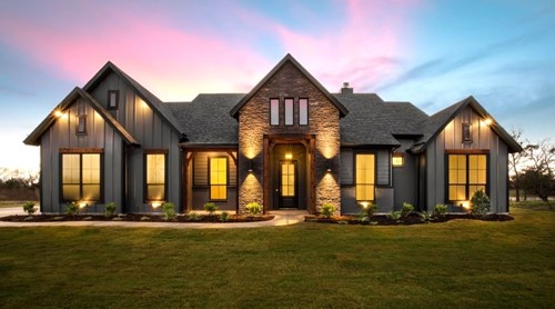 Large house with stone and grey siding, illuminated by roofline spotlights and two large sconces by the grand front door, captured at dusk