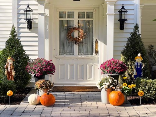 All-white traditional-style home with front porch decorated with purple mums, pumpkins, and small scarecrows on a bright sunny day