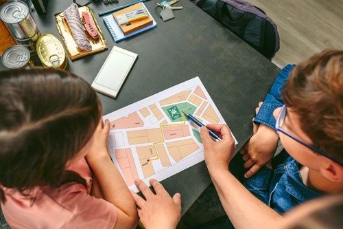 Over-the-shoulder view of a parent showing their son and daughter a neighborhood map, with emergency provisions on the table in the background