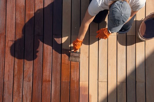 Aerial view of a man in a ball cap and overalls sealing a wooden deck with a brush, halfway through the job, with a can of sealant beside him