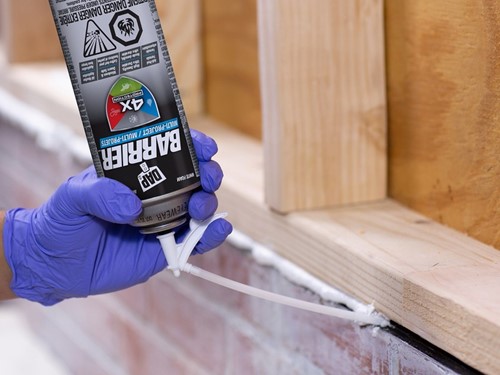 Gloved hand applying barrier straw foam sealant to the gap between brick and wood framing, with the can held upside down