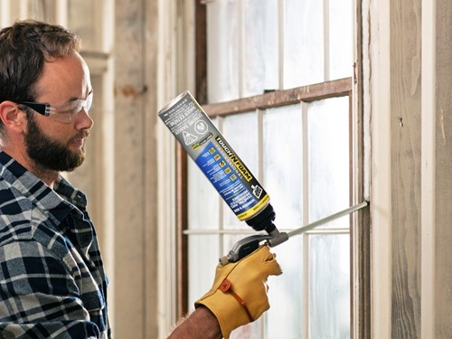 Man applying gun foam sealant to the gap around a window casing, holding the canister upside down while wearing protective gloves and glasses