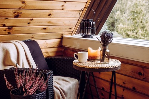 Black wicker chair with a cozy beige blanket, next to a small table holding a steaming cup of tea and a candle, angled to look out the window in a wood-paneled room