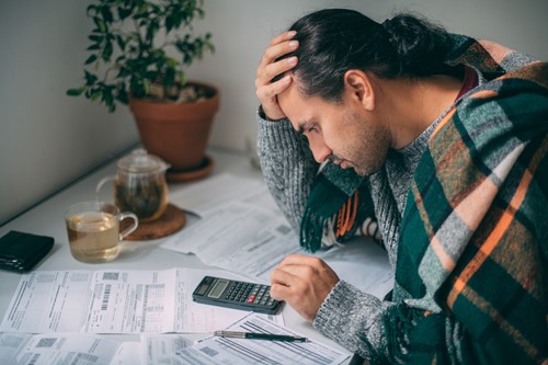 Man wrapped in a plaid blanket, leaning over a table with a pile of bills and a calculator, with a cup of tea nearby.