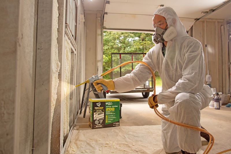 Man in a white protective suit and respirator applying spray foam sealant to a wall.