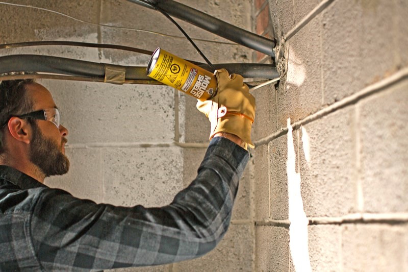 Man wearing safety glasses and a yellow work glove spraying MOUSE SHIELD foam sealant into a pipe penetration on a cinderblock wall.