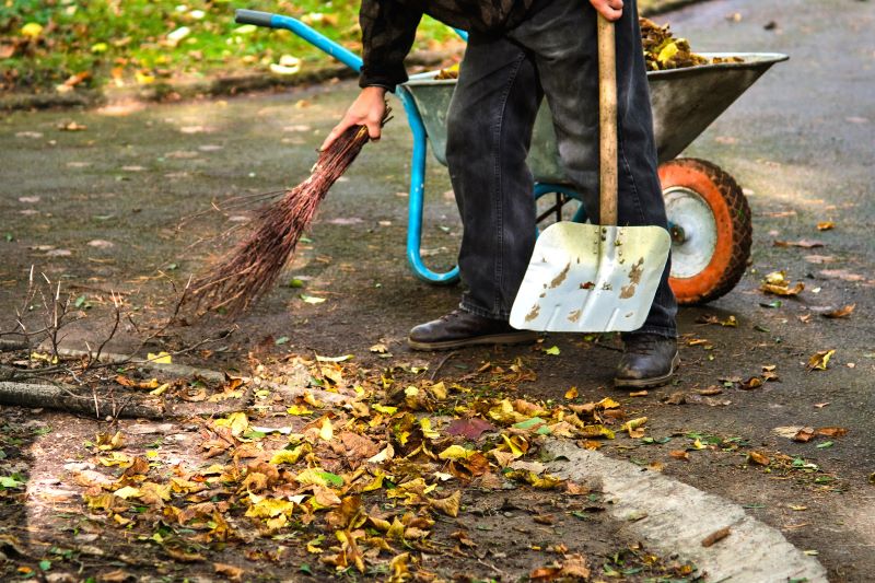 Man holding a bundle of twigs and a shovel, cleaning fallen leaves from the ground, with a wheelbarrow filled with leaves behind him.