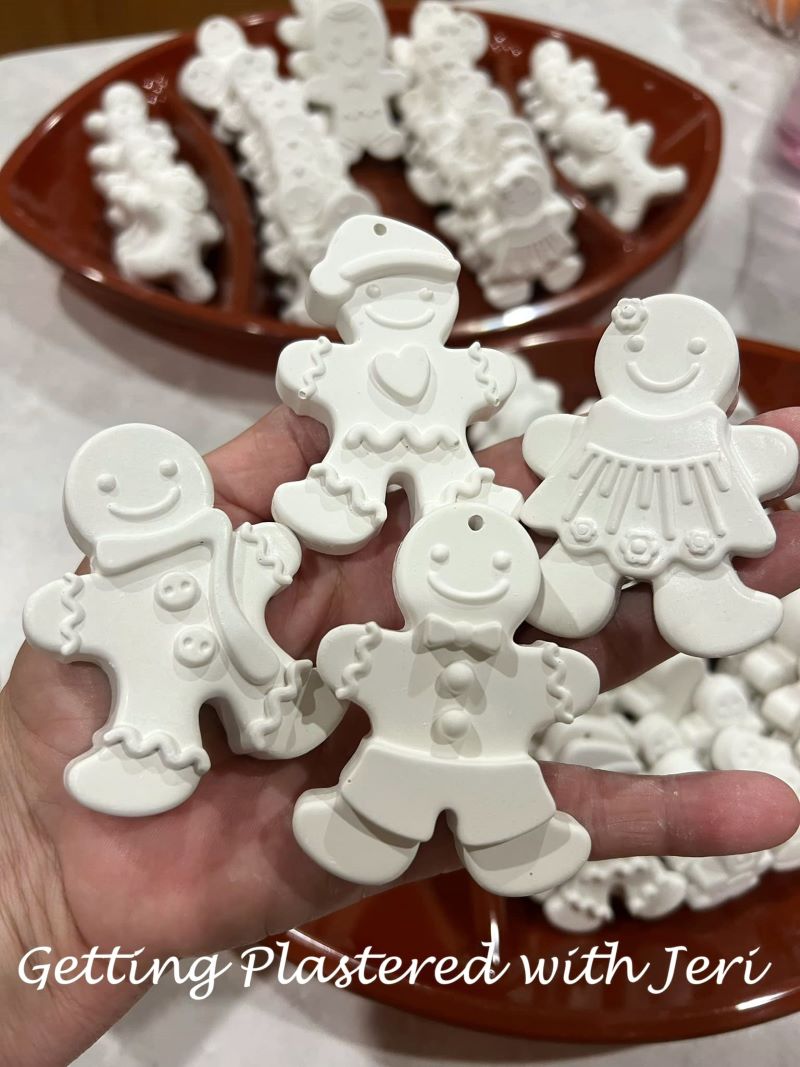 Handmade plaster ornaments shaped like gingerbread figures, shown before being painted. The ornaments are displayed in a hand with more ornaments in the background.