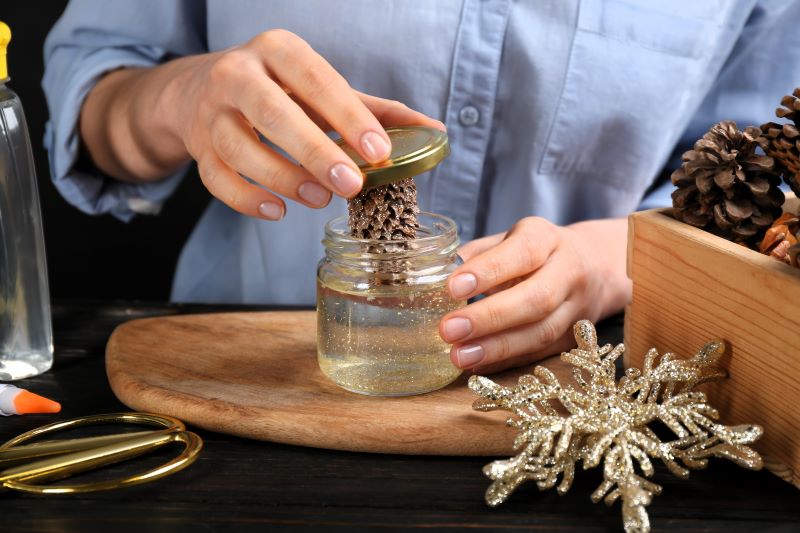 A person assembling a DIY snow globe by placing a pinecone attached to a jar lid into a glass jar filled with water and glitter.