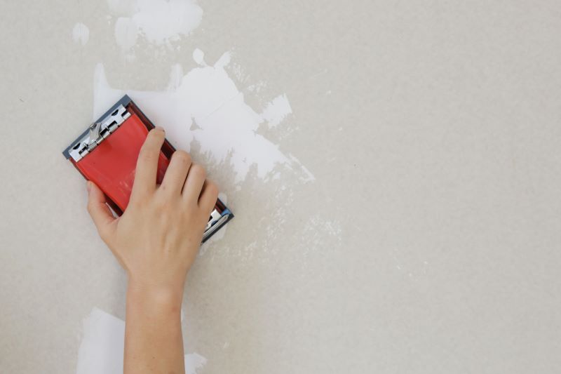 A person sanding a white patch on a wall with a red sanding block, preparing the surface for painting.