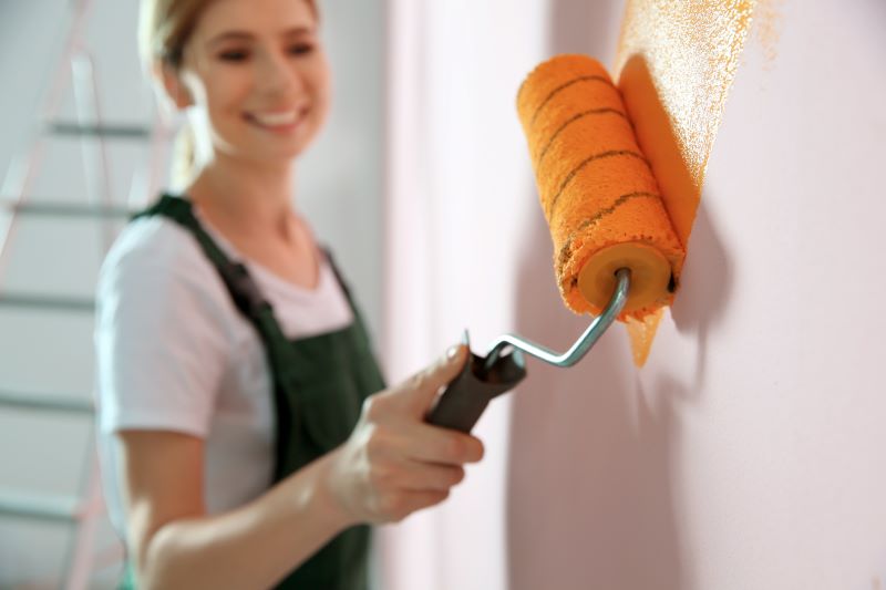 A smiling person using an orange paint roller to apply fresh paint to a wall, with a ladder visible in the background.